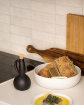 The same beige stoneware shallow serving bowl now filled with bread, placed beside a matching olive oil dispenser and a small flat plate holding a side dish, all arranged on a simple, neutral-toned surface.